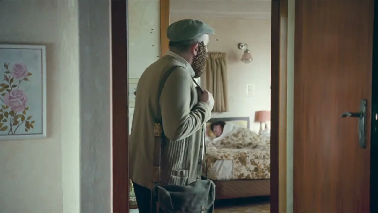 A man with a swarm of bees on his face walks past a bedroom where another man is sleeping, seen through a doorway.