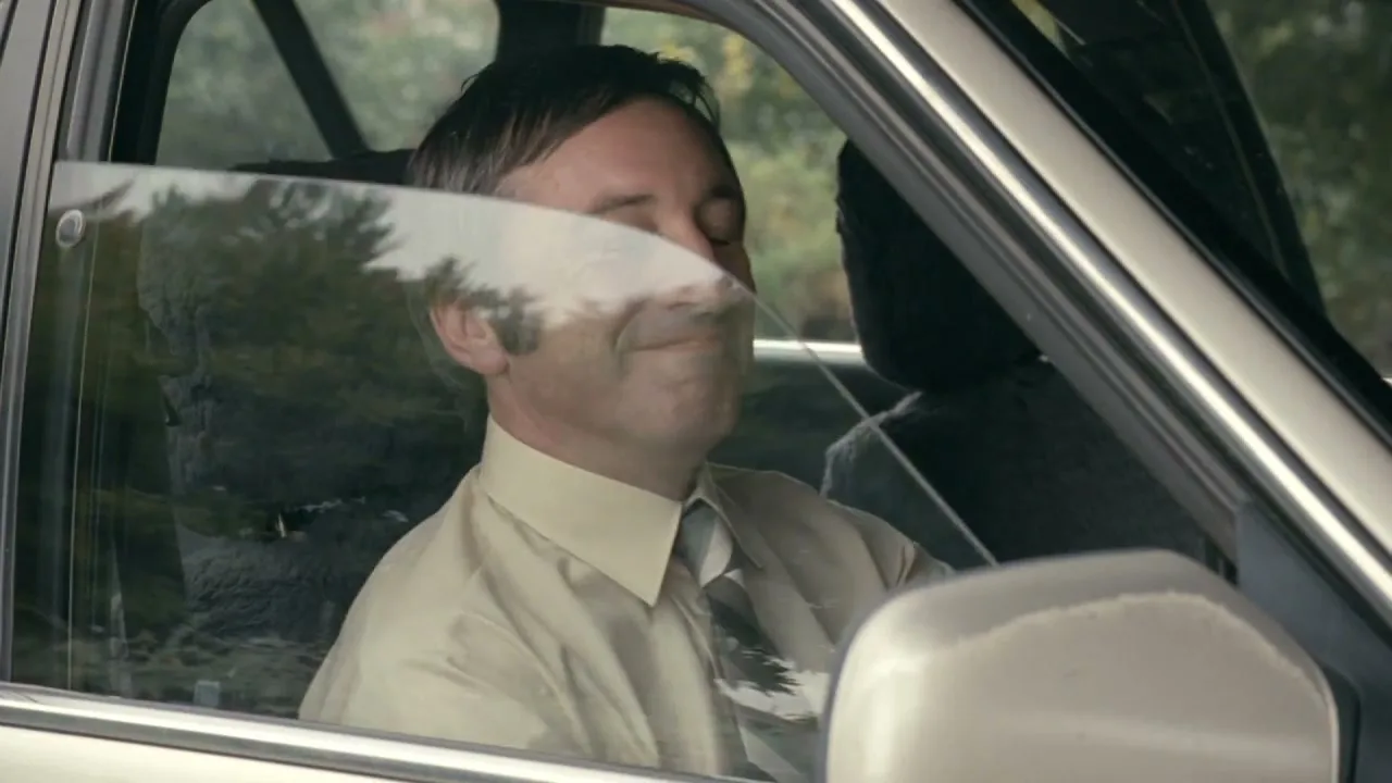 A middle-aged man in a shirt and tie is driving a car, looking out the window and smiling.