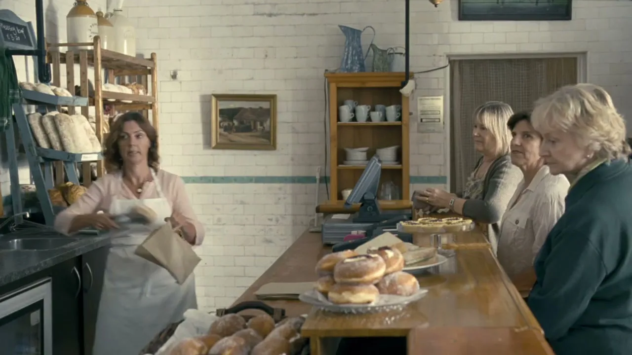 A female baker in an apron serves three older women waiting in line at a rustic bakery.