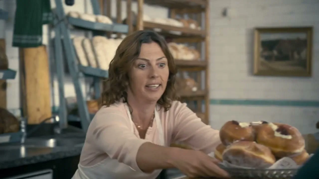 A female baker with brown hair and a pink shirt, wearing an apron, looks surprised as she hands a plate of donuts to a customer in a rustic bakery.