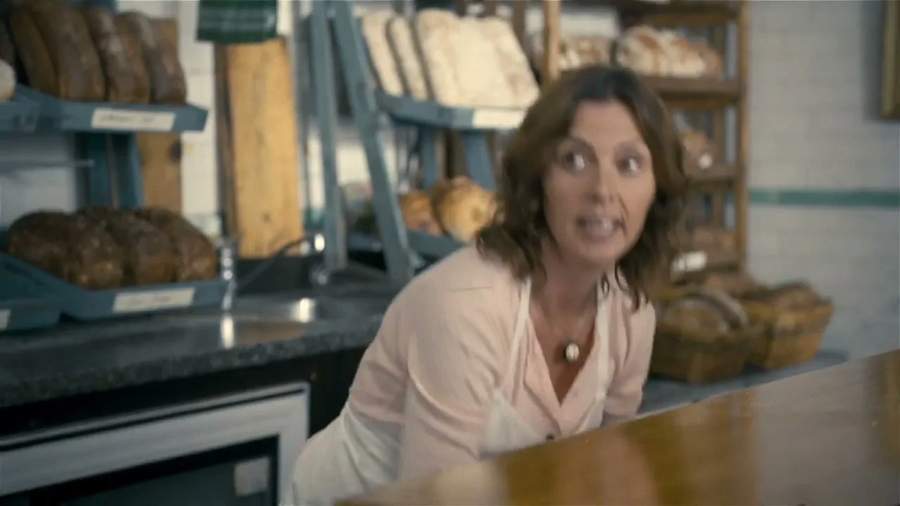 A female baker with brown hair and a pink shirt smiles as she leans over a counter in a rustic bakery.