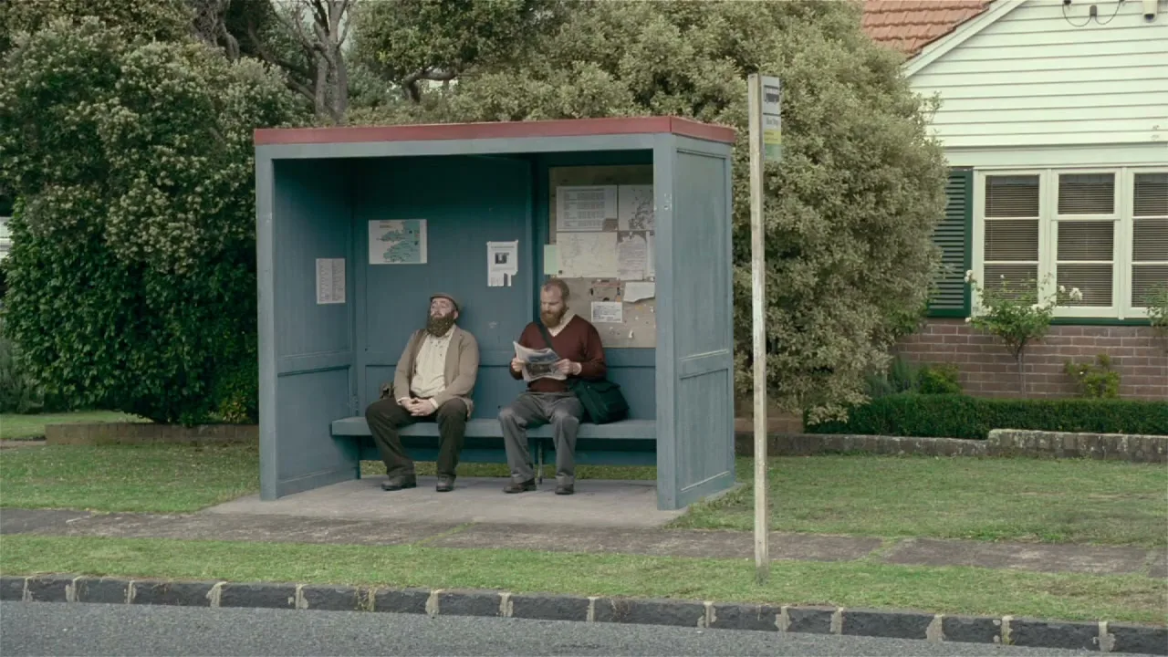 Two bearded men sit at a bus stop, one reading a newspaper, in a suburban neighborhood on a bright day.