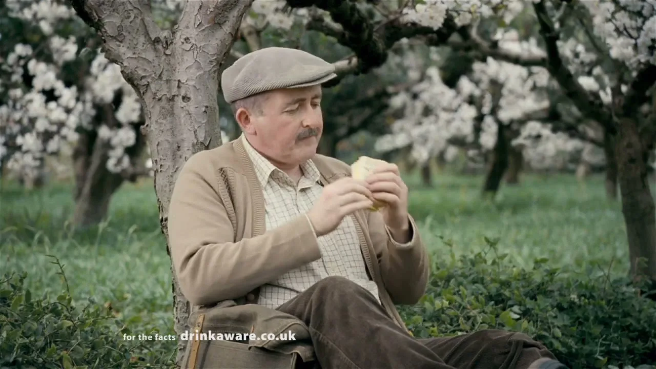 A man in a flat cap and cardigan sits under a blossoming tree in an orchard, eating a sandwich.