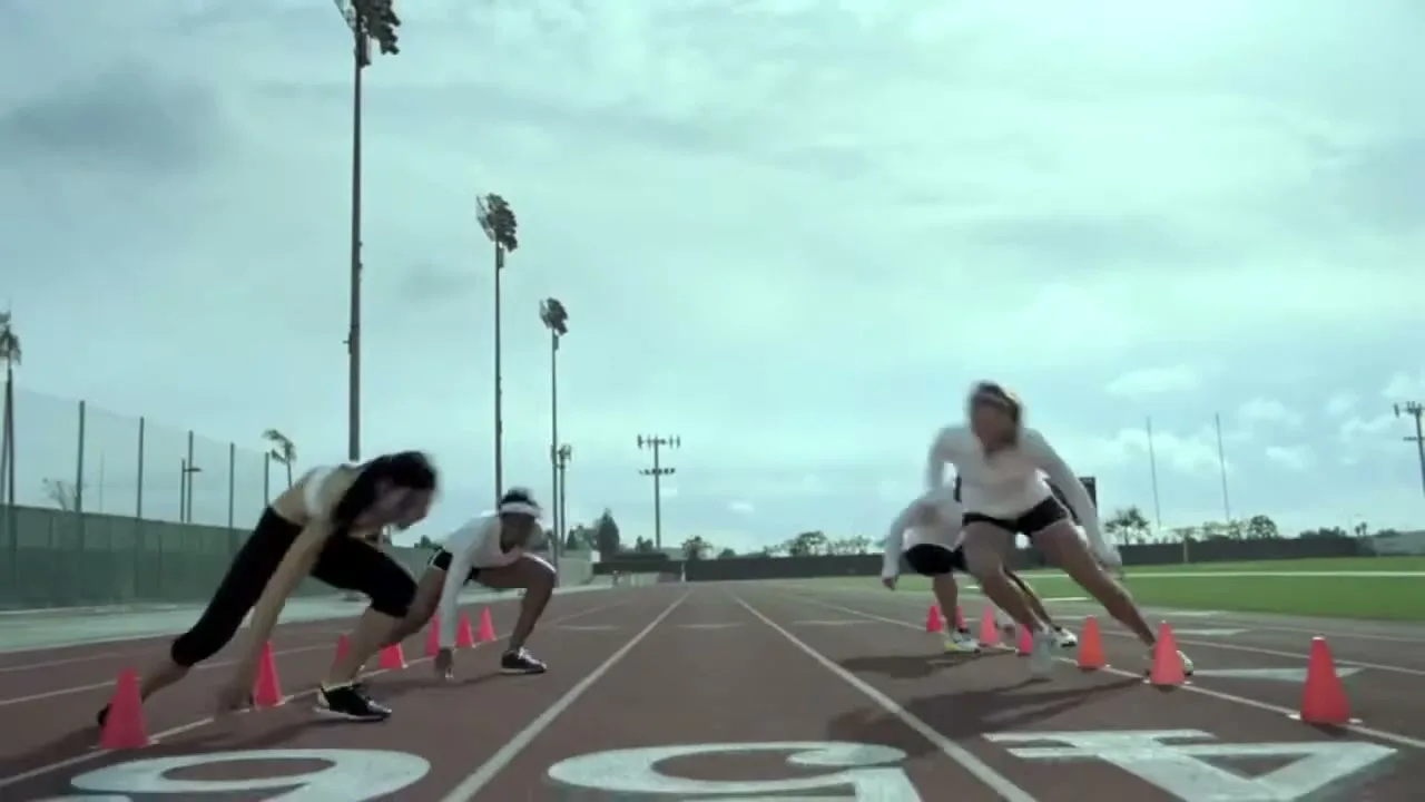 Four female athletes in white tops and black shorts/pants run a cone drill on a track and field during the day.