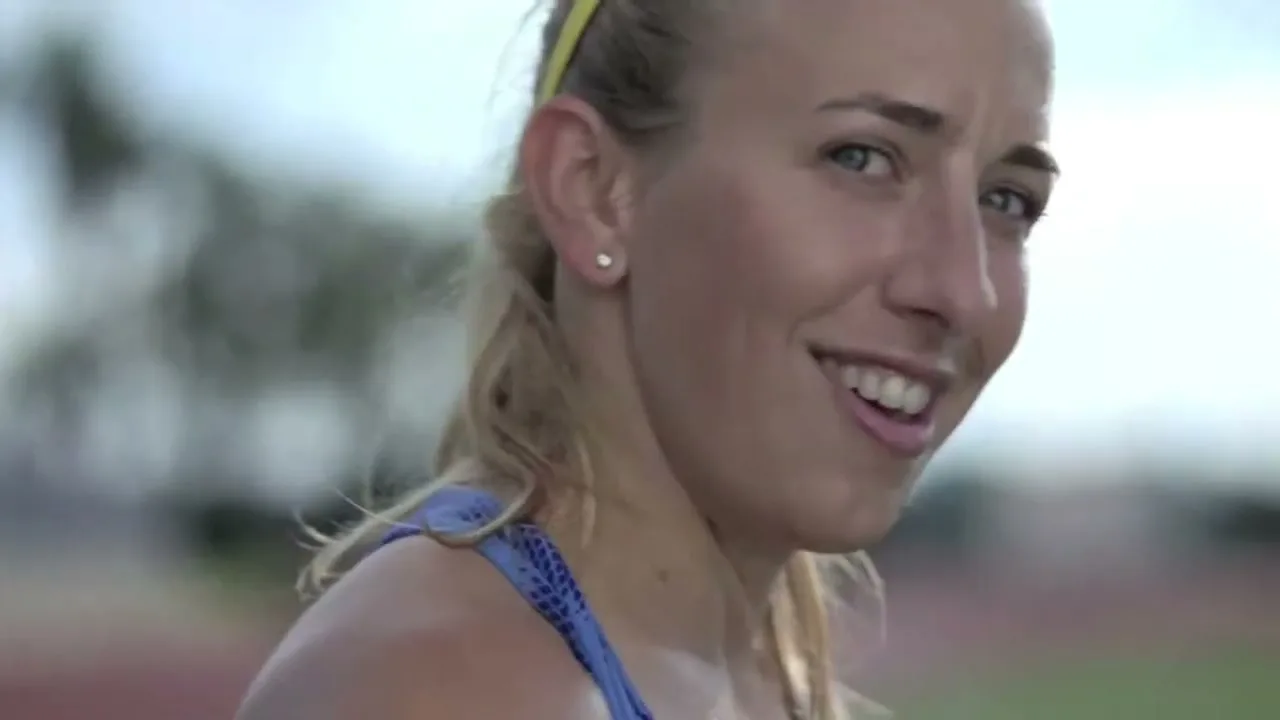 A female athlete with blonde hair and a yellow headband smiles and looks at the camera on a track field.