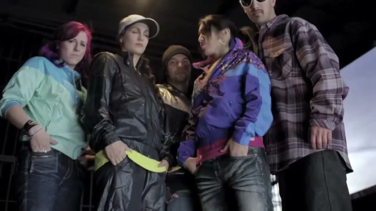 A diverse group of five young adults, dressed in casual streetwear, stand together looking down at the camera with serious expressions in an urban setting.