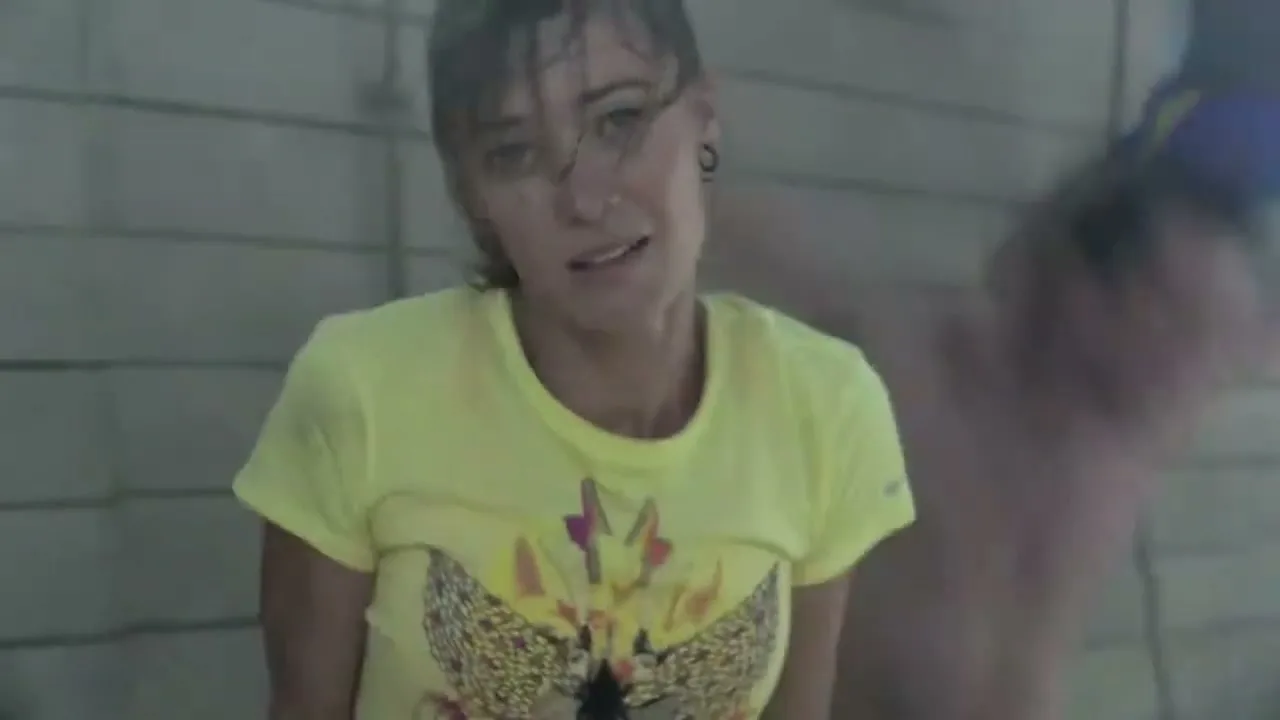 A female athlete in a yellow t-shirt with wet hair looks distressed while being held by another person in a locker room.