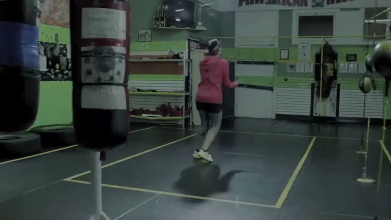 A female boxer in a pink hoodie and grey leggings jumps rope in a boxing gym, with punching bags in the foreground and background.
