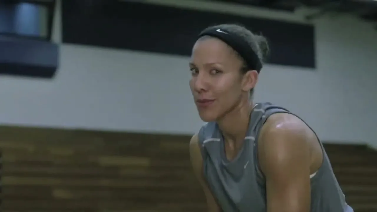 A female basketball player in a grey tank top and black headband looks at the camera, then turns to her left, on a basketball court.