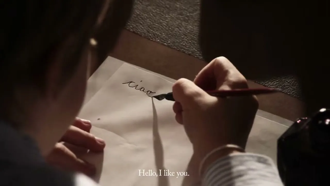 A young person with light hair writes the word "ciao" on a piece of paper with an ink pen, seen from an overhead perspective.