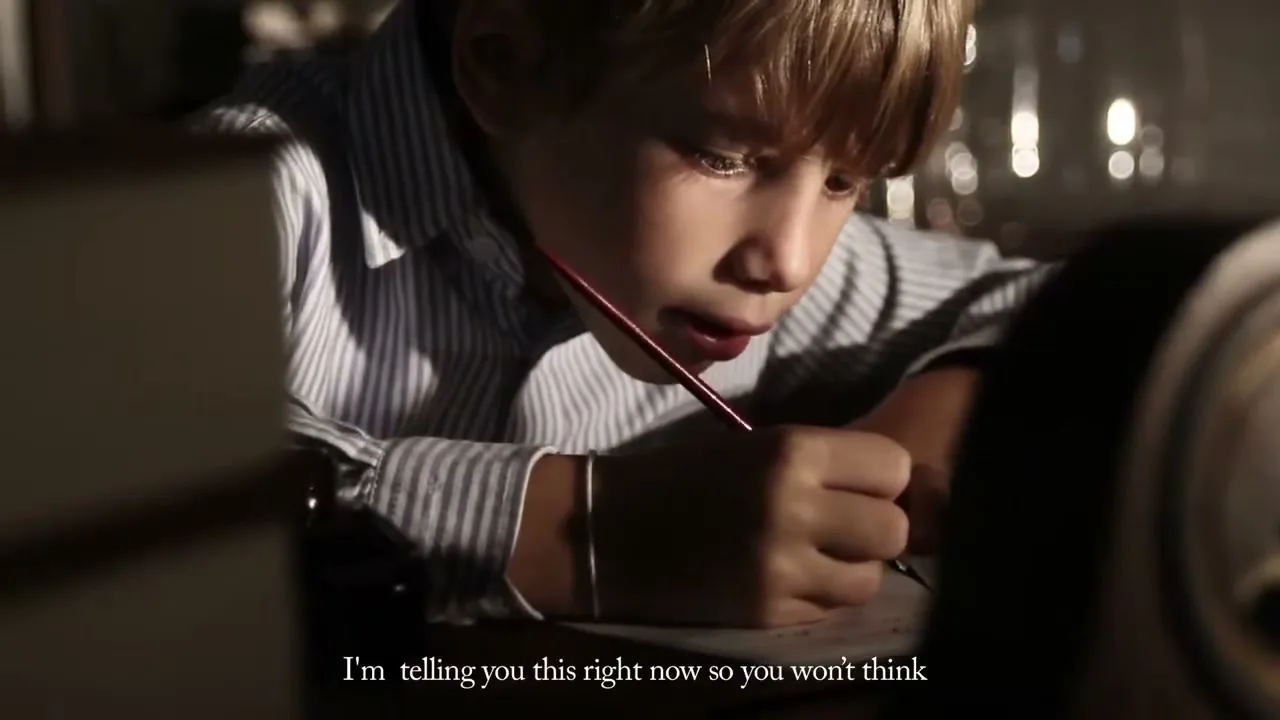 A young boy with blonde hair, wearing a striped shirt, intently writes a letter with a pen at a desk in a dimly lit room.
