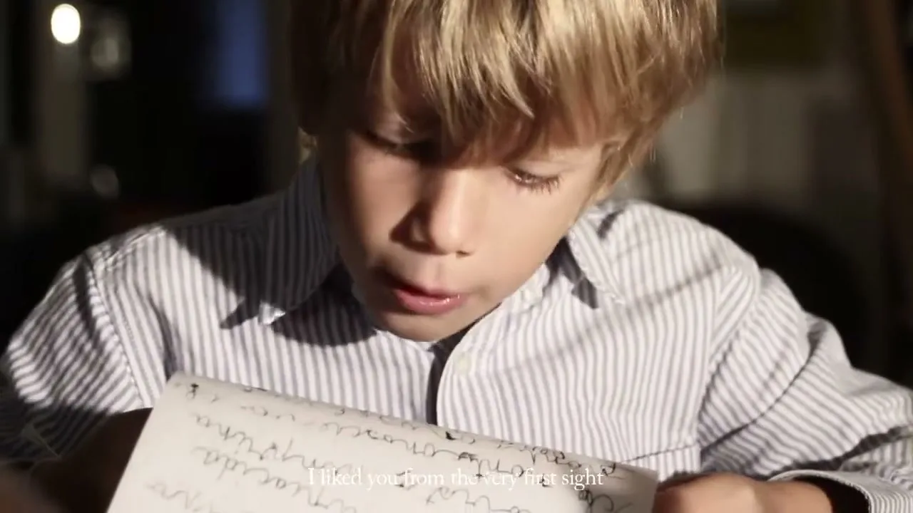 A young boy with blonde hair and a striped shirt reads a handwritten letter, looking surprised and then thoughtful in a dimly lit interior.