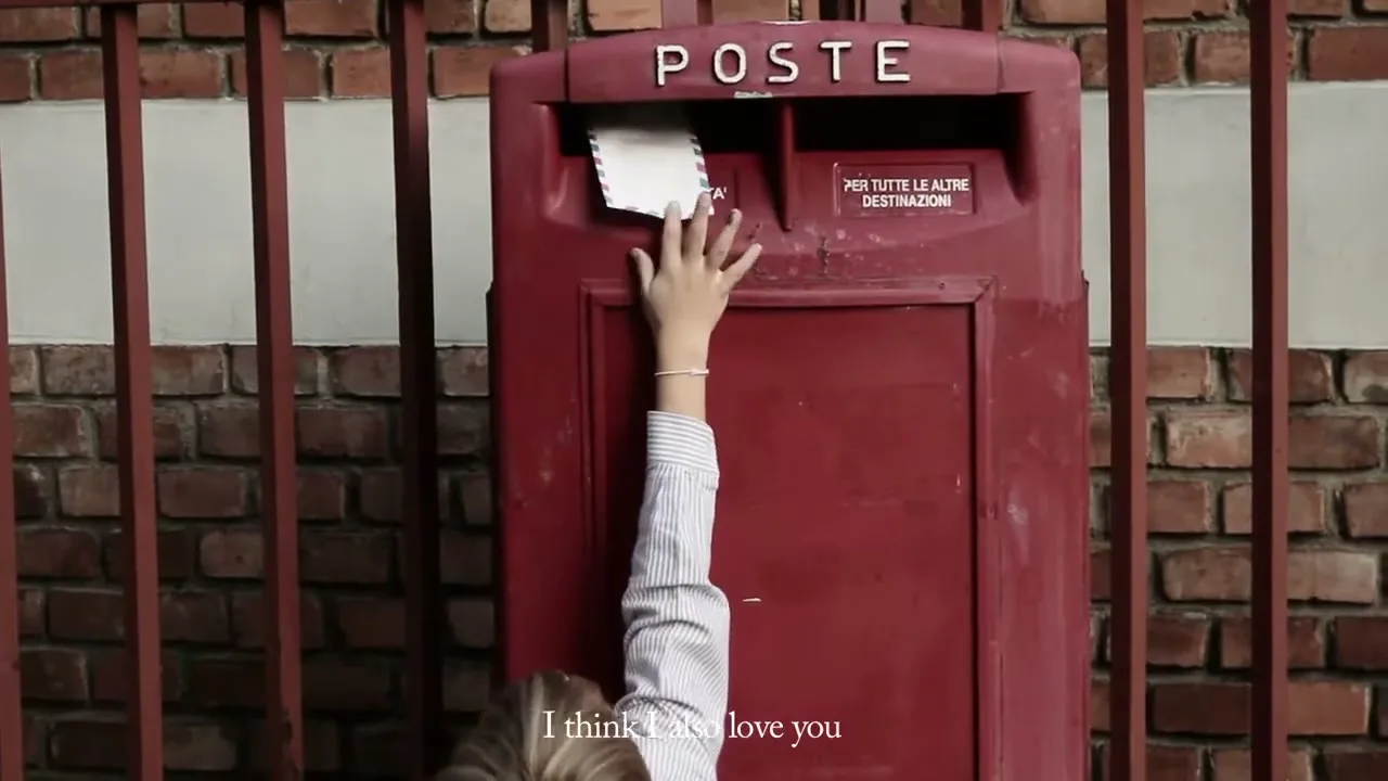 A young girl in a striped shirt reaches up to put a letter into a red Italian mailbox against a brick wall.