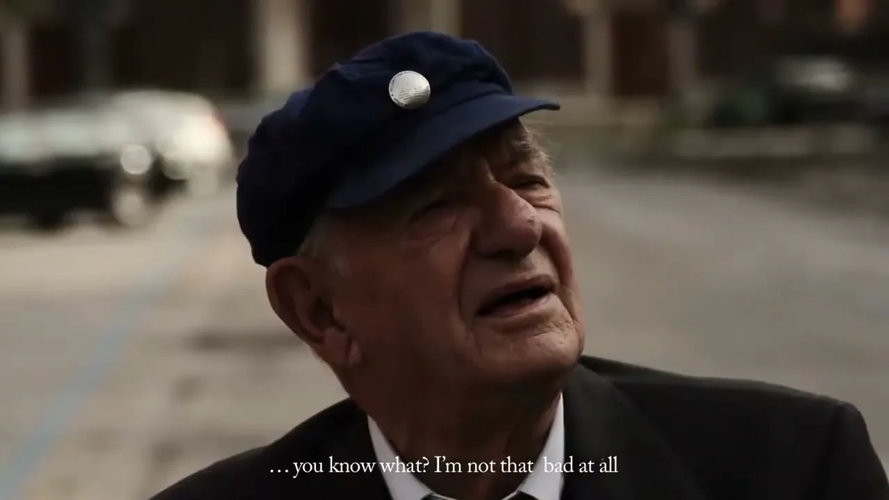 An elderly man in a dark suit and blue cap looks up and to the right, with a car visible in the blurry background.
