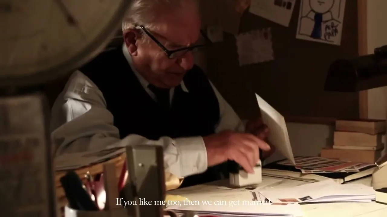 An elderly man with glasses and a vest stamps letters at a cluttered desk in a dimly lit office.