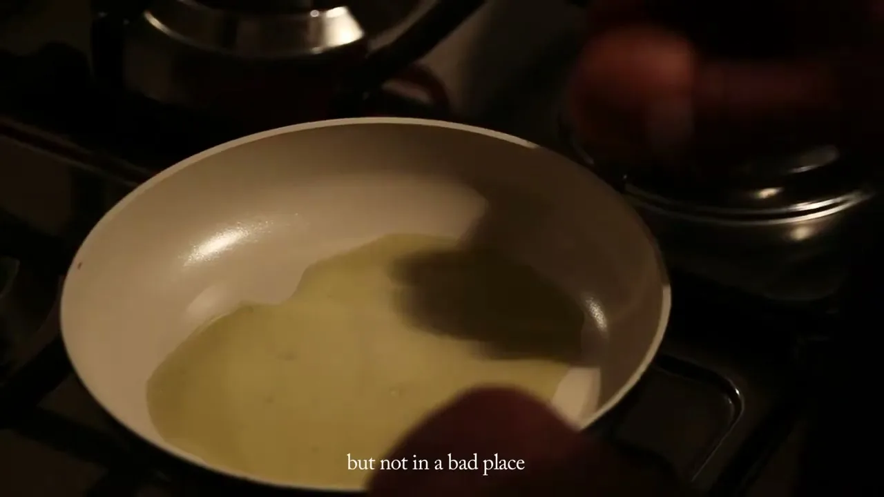 A person's hands hold an egg over a frying pan with oil, preparing to cook in a dimly lit kitchen.