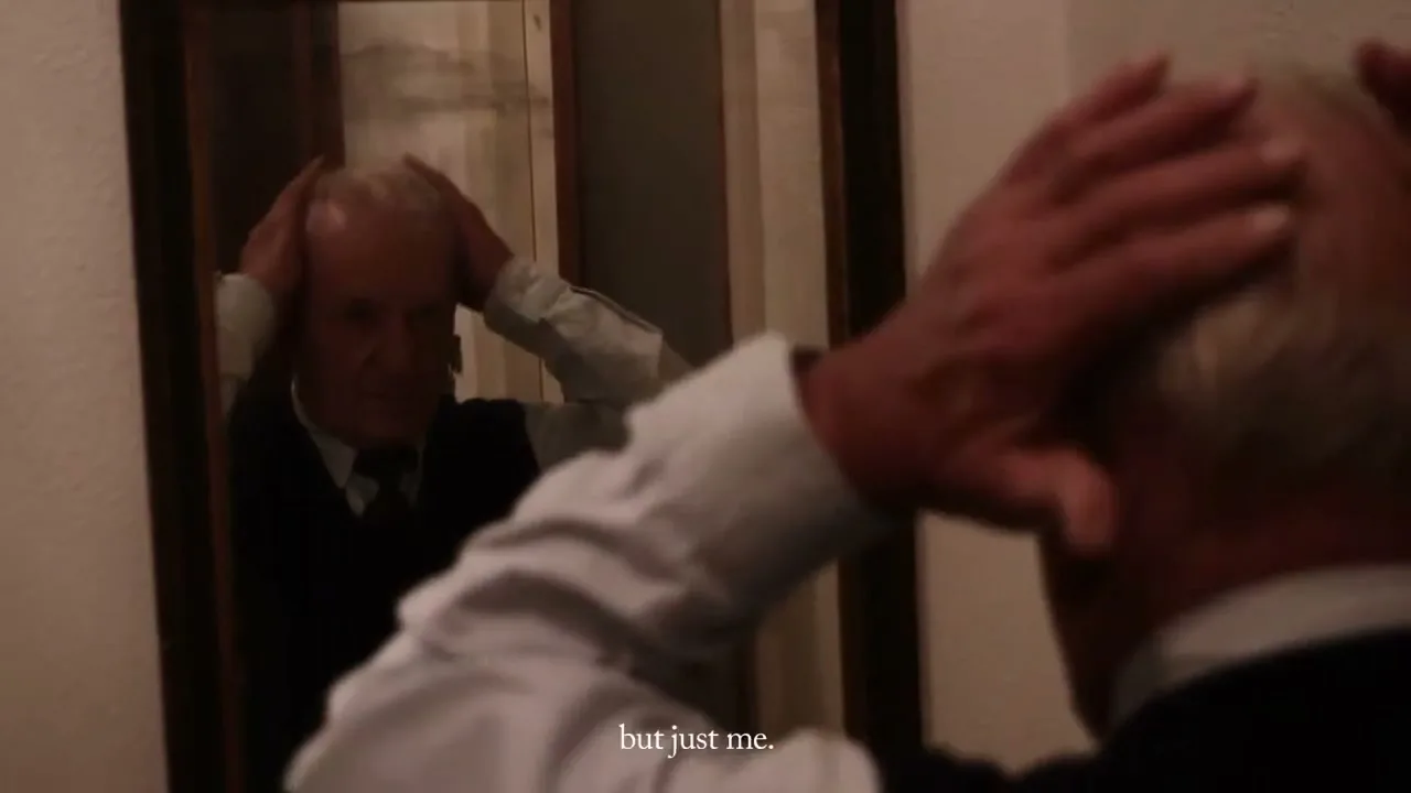 An older man with white hair, wearing a white shirt and dark vest, looks at his reflection in a mirror, adjusting his hair and holding red roses in an interior setting.