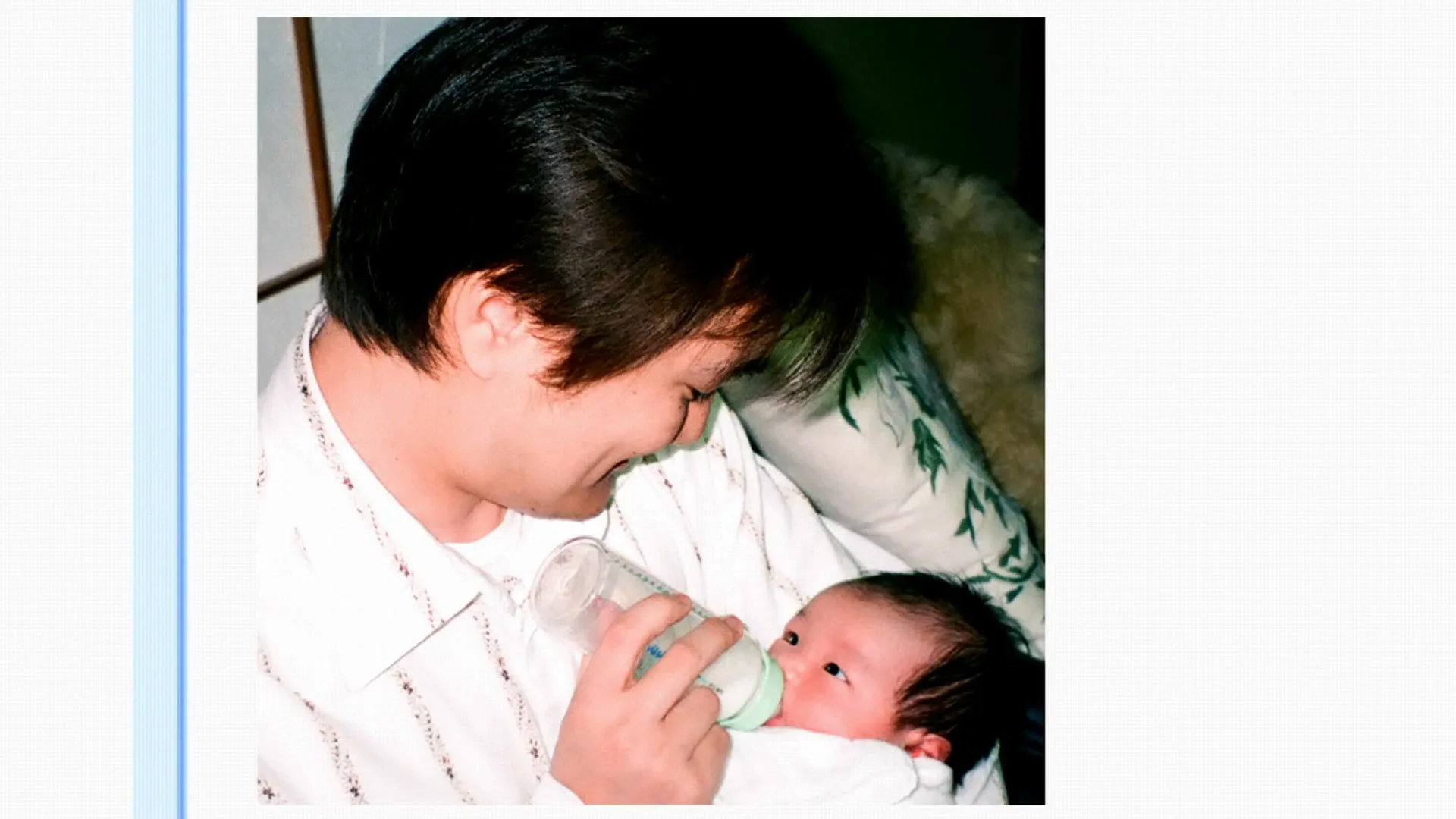 A father with dark hair and a white shirt smiles at his baby while feeding it a bottle in a warm, natural light setting.
