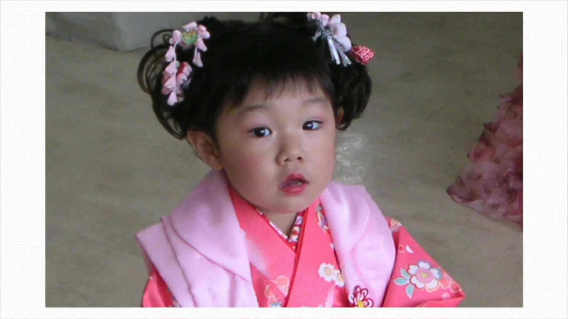 A young Asian girl with dark curly hair, wearing a pink kimono and hair ornaments, looks up with a slightly open mouth.