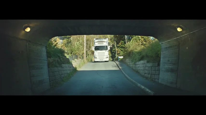 A white truck emerges from a tunnel on a sunny day, driving towards the camera on a paved road surrounded by green foliage.