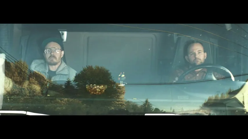 Two male truck drivers are seen through the windshield of their truck, with reflections of trees and power lines on the glass, as they drive down a road.