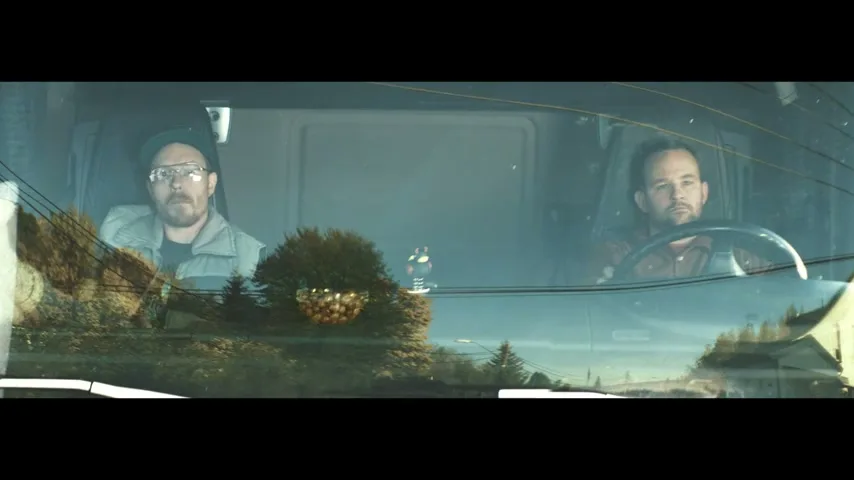Two male truck drivers, one wearing glasses and a cap, the other with a beard, are seen through the windshield of their truck, driving on a road with trees and power lines reflected on the glass.