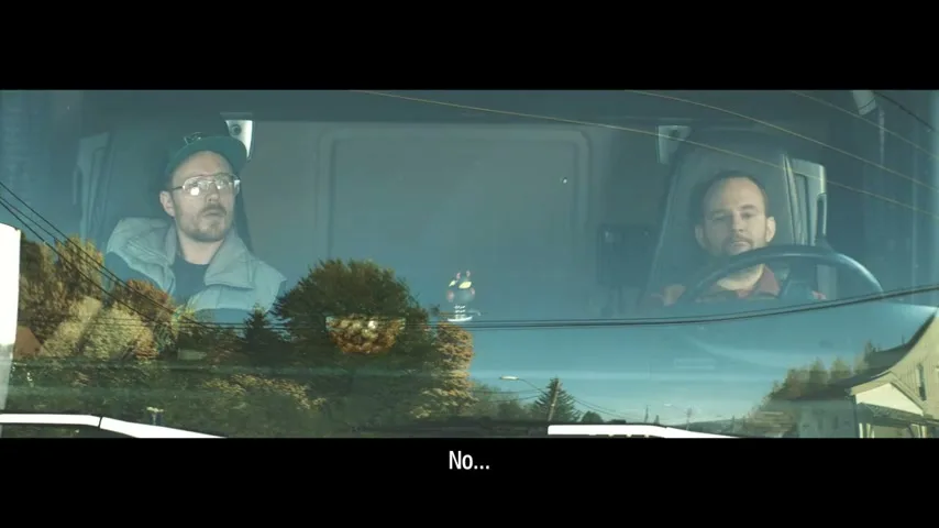 Two men in a truck, one driving and one in the passenger seat, are seen through the windshield, with reflections of trees and houses.
