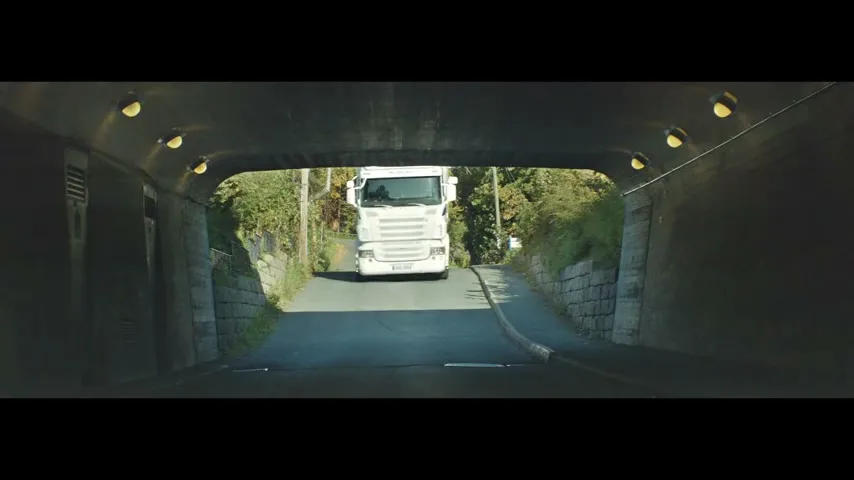 A white truck emerges from a tunnel on a sunny day, driving towards the camera on a winding road surrounded by green trees.
