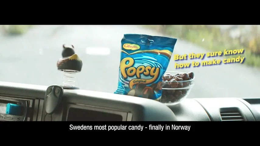 A bag of Cloetta Popsy candy and a bowl of chocolate balls sit on a truck dashboard, with a bobblehead toy and a road visible through the windshield.