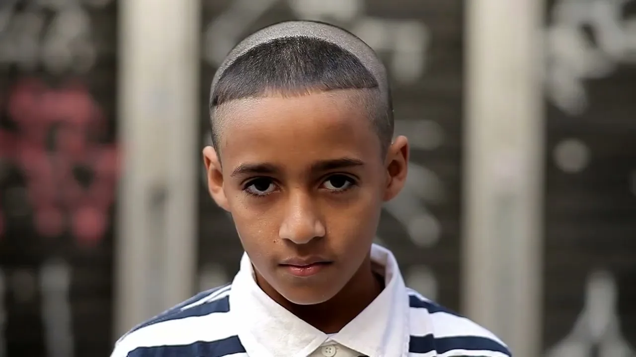 A young boy with a fresh haircut and striped shirt looks directly at the camera in an urban setting.