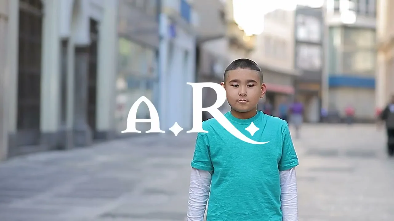 A young boy with a buzz cut and a teal shirt stands with his eyes closed, then opens them, on a city street with a large white graphic overlay.