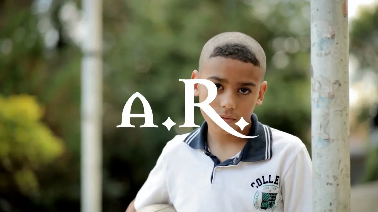 A young boy with a unique haircut and a white polo shirt stands outdoors, holding a soccer ball, looking directly at the camera.