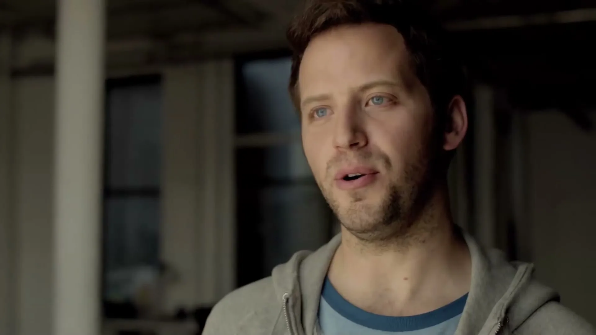 A man with blue eyes and stubble, wearing a grey hoodie and blue t-shirt, looks off-camera and speaks in an interior setting.