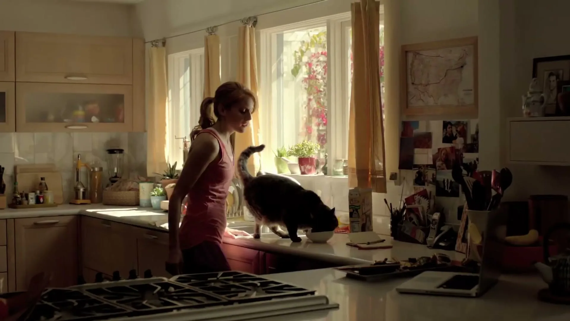 A young woman with red hair in a pink tank top and shorts pets a dark cat on a kitchen counter in a sunlit kitchen.