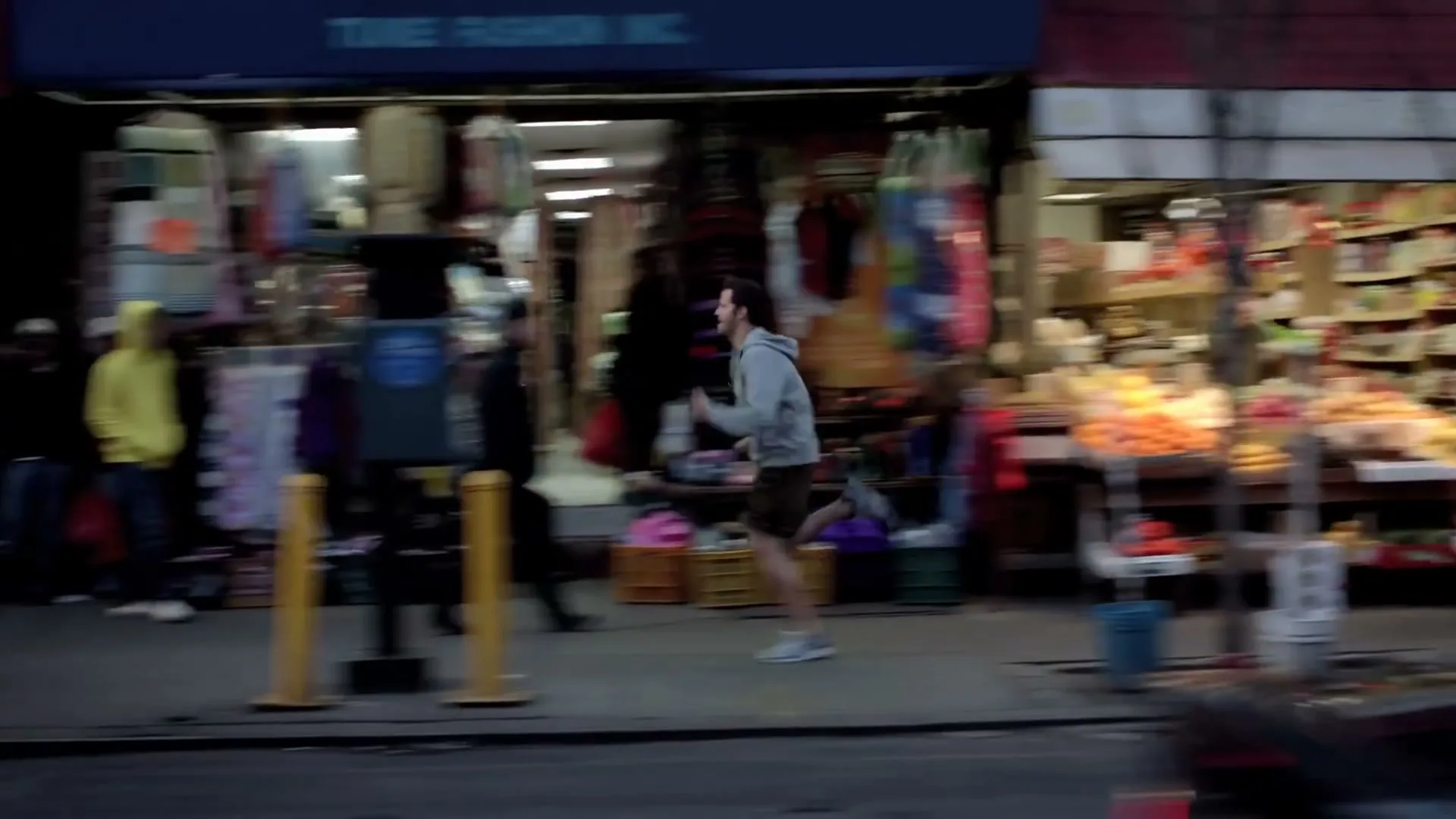 A man in a grey hoodie and shorts runs past storefronts on a busy city street, with blurred cars in the foreground.