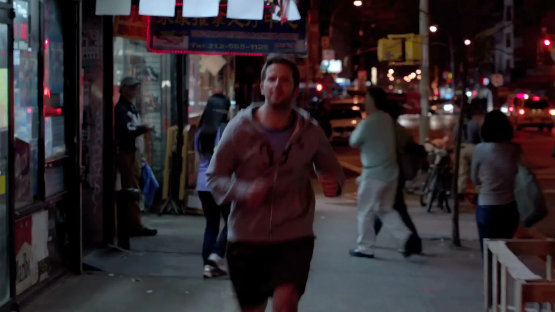 A man in his 30s, wearing a grey hoodie and shorts, runs down a busy city sidewalk at night, looking determined.