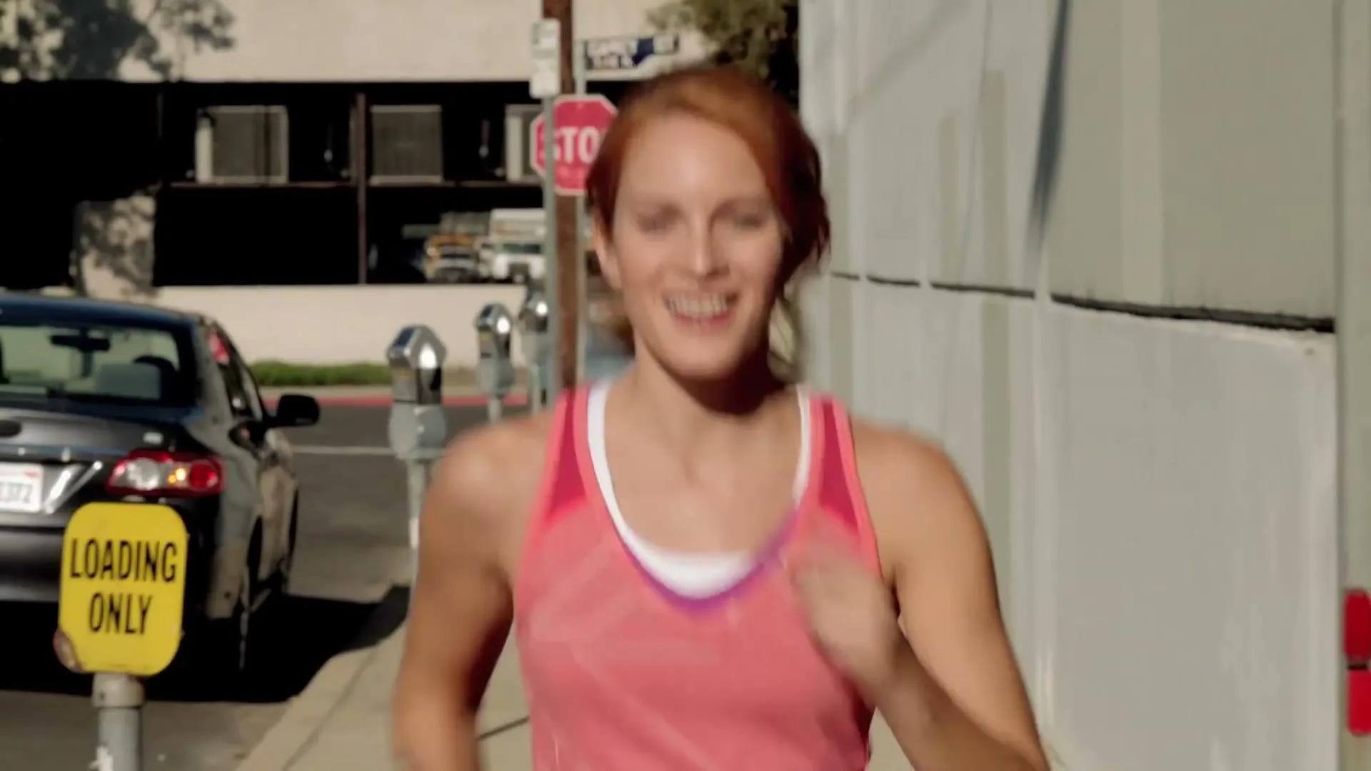 A red-haired woman in a pink tank top runs on a sunny sidewalk, smiling as she passes a stop sign and parked cars.