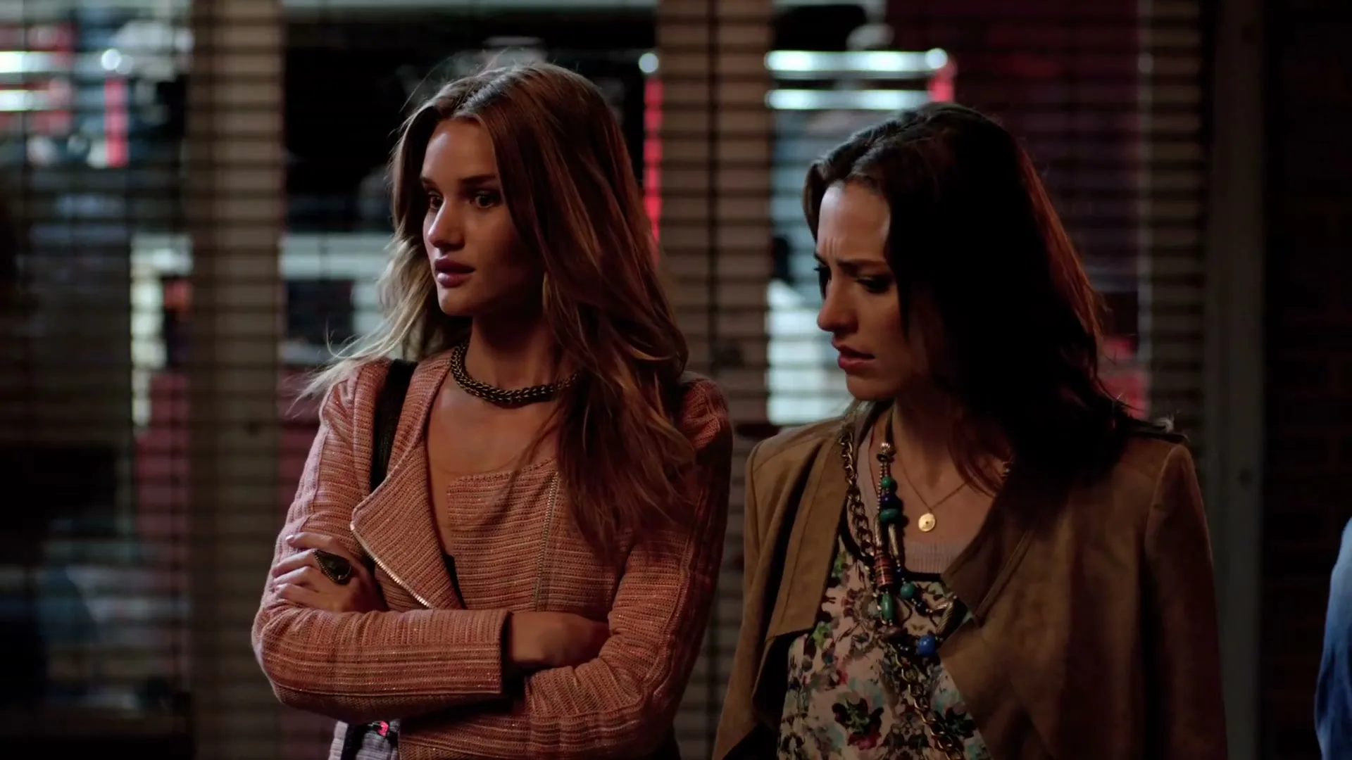 Two women, one with long blonde hair in a pink jacket and the other with dark hair in a brown jacket, stand looking concerned on a city street at night.
