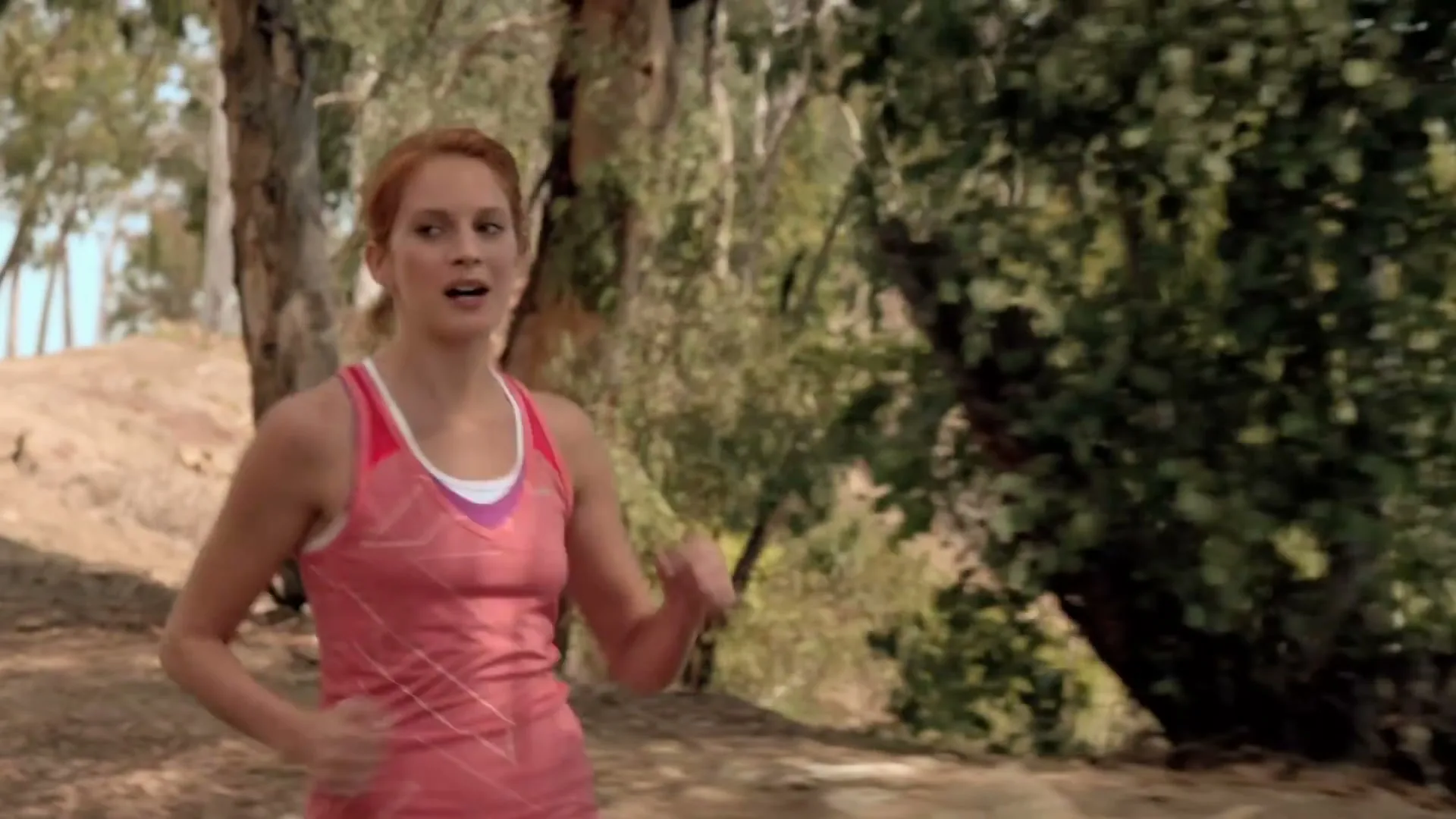 A young woman with red hair, wearing a pink tank top, runs on a dirt path through a wooded area during the day.