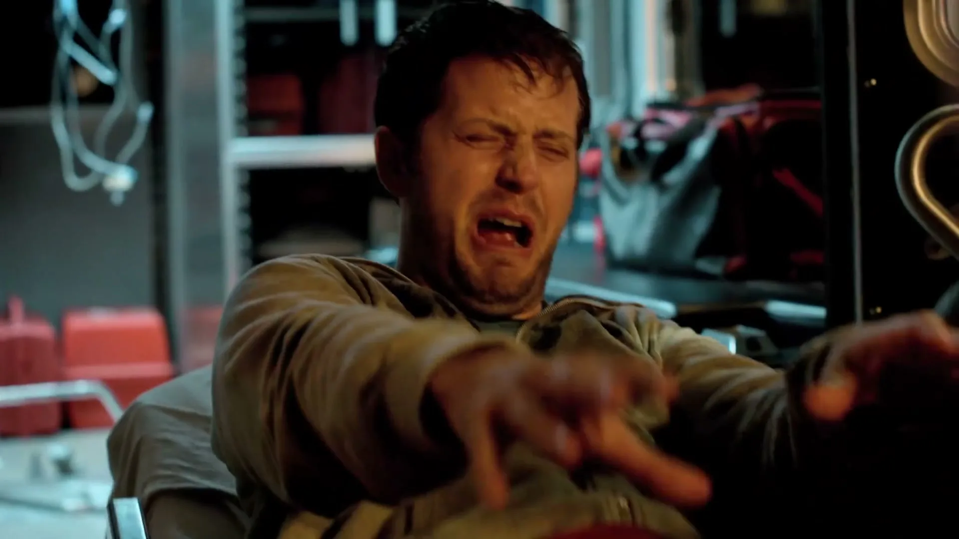 A man in his 30s, wearing a grey hoodie, screams in pain while sitting in an ambulance.