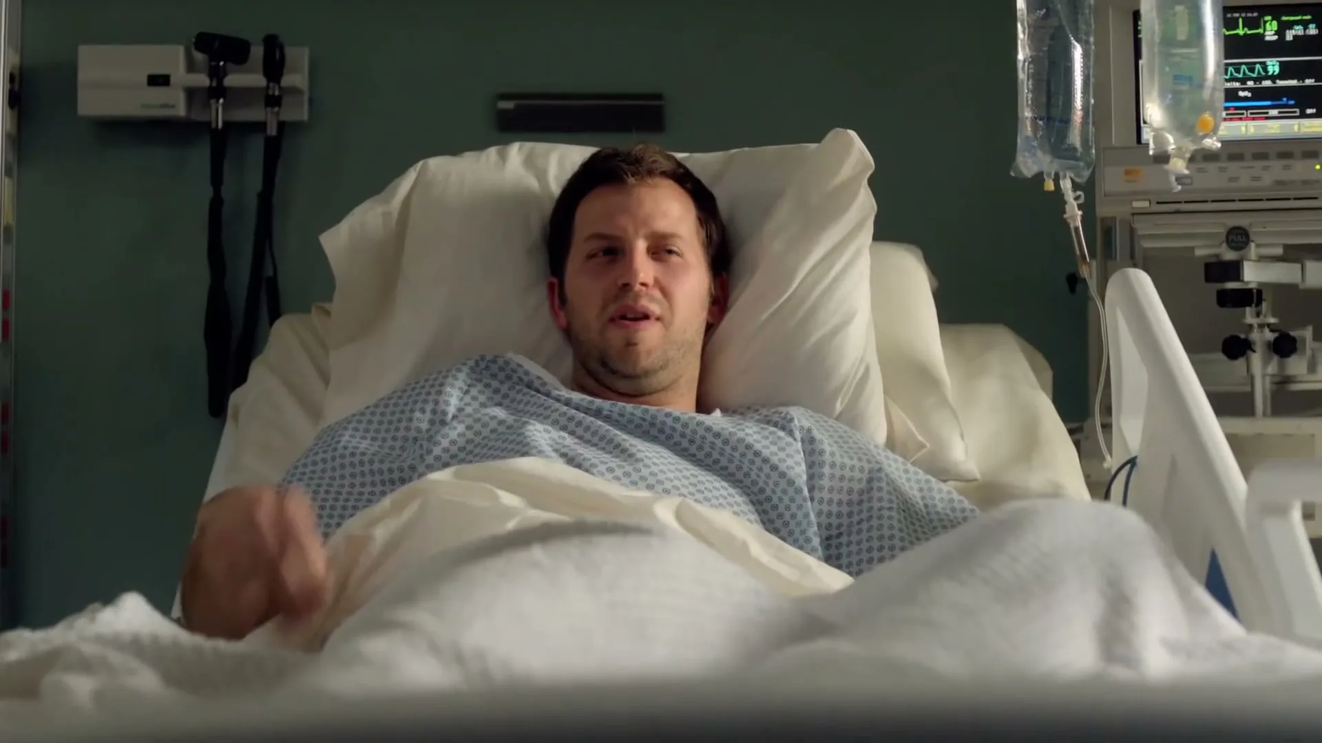 A male patient in his 30s lies in a hospital bed, looking distressed and clutching his chest in an interior hospital room.