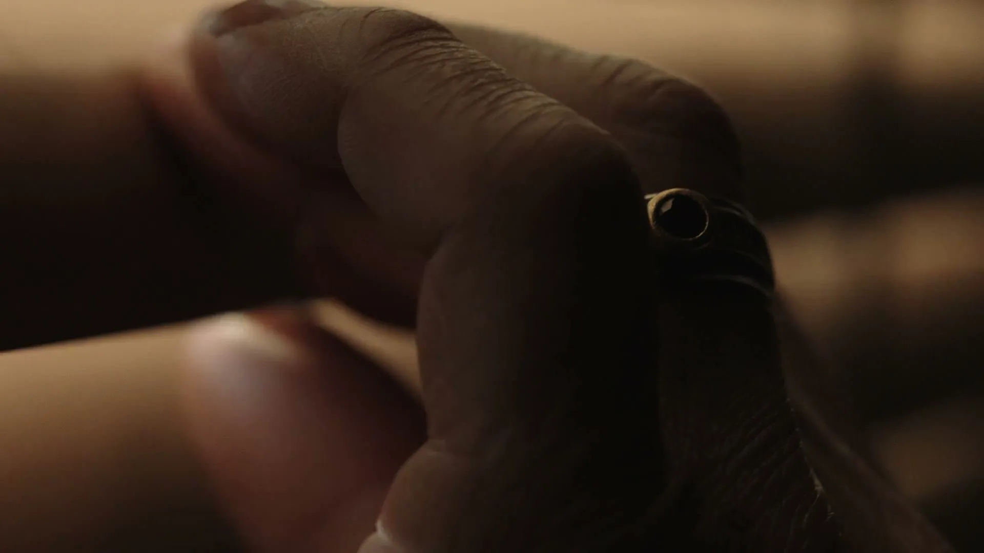 A person's hand with a ring on the finger is shown in a dark, intimate setting, with soft light illuminating the skin.