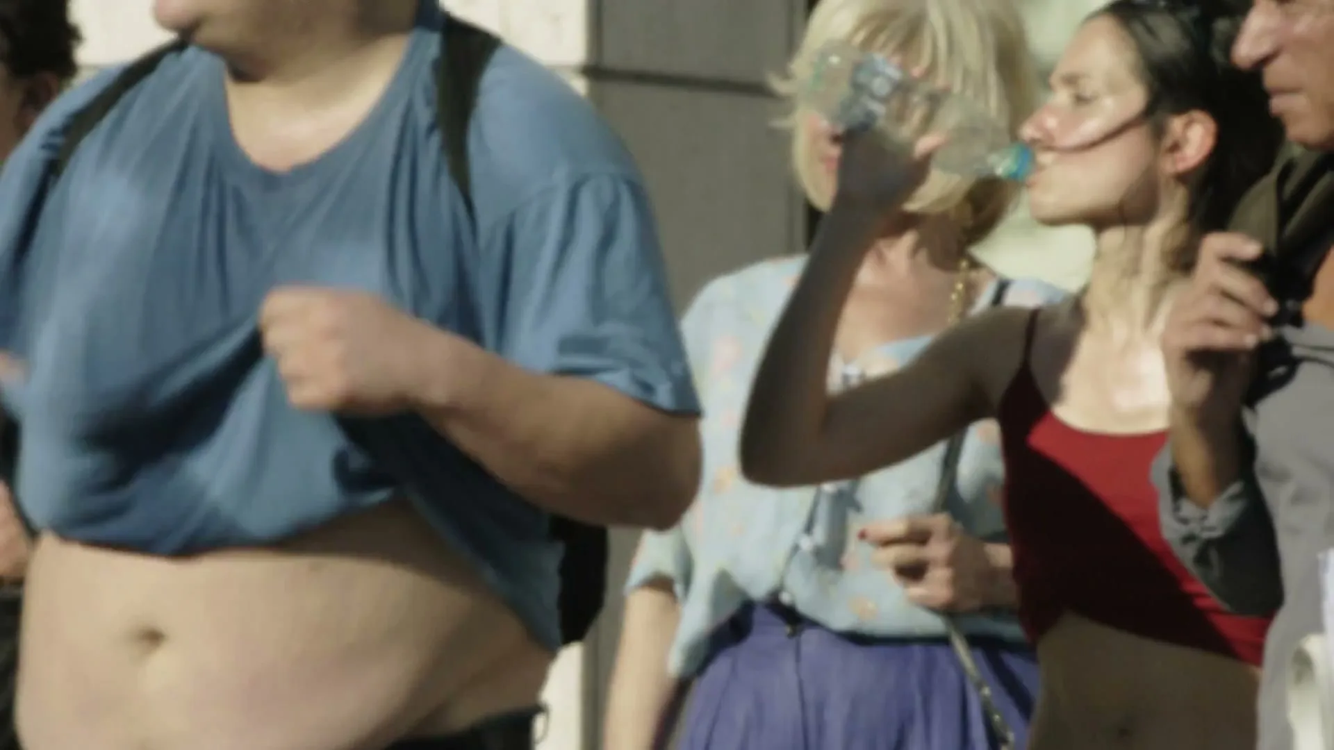 A man lifts his blue t-shirt to reveal his sweaty stomach, while a woman next to him drinks water from a plastic bottle on a sunny day.
