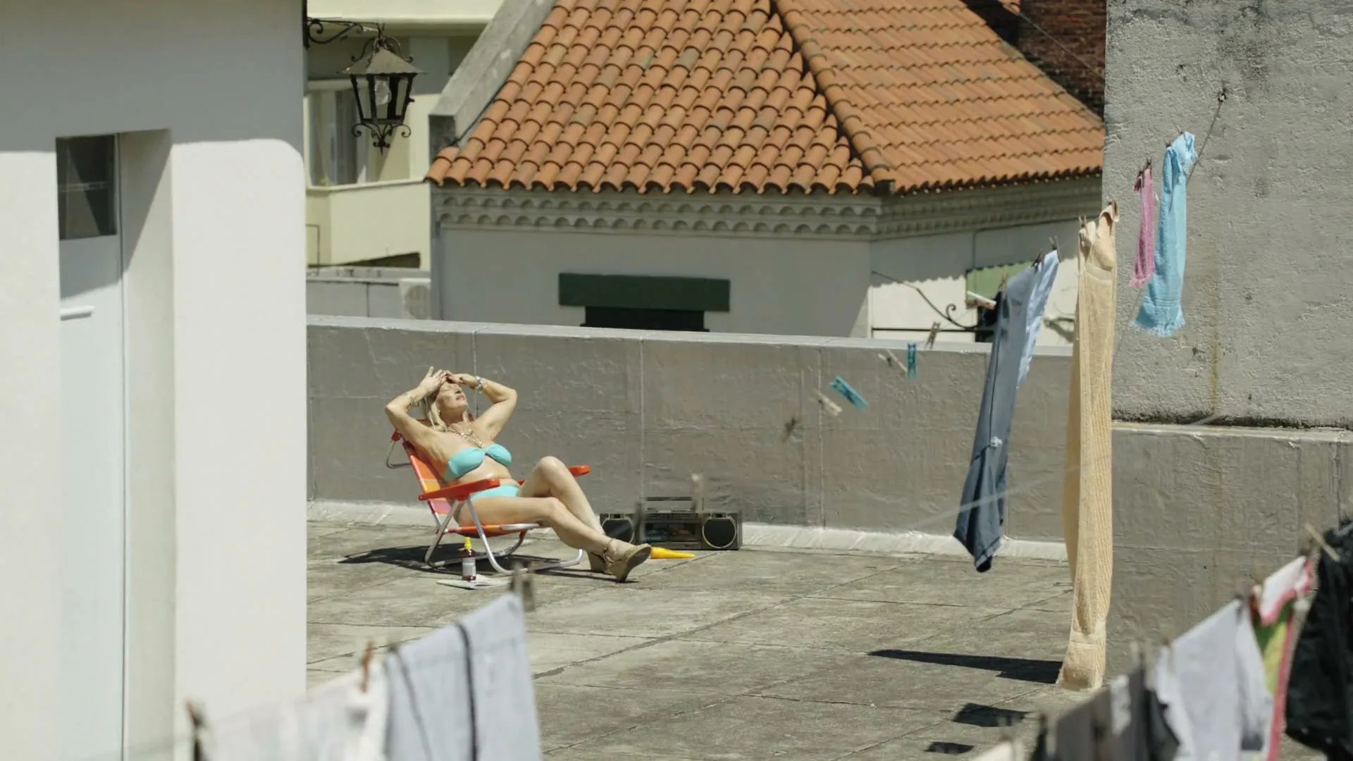 A woman in a bikini sunbathes on a rooftop next to a boombox, with laundry hanging on lines in the foreground and background.