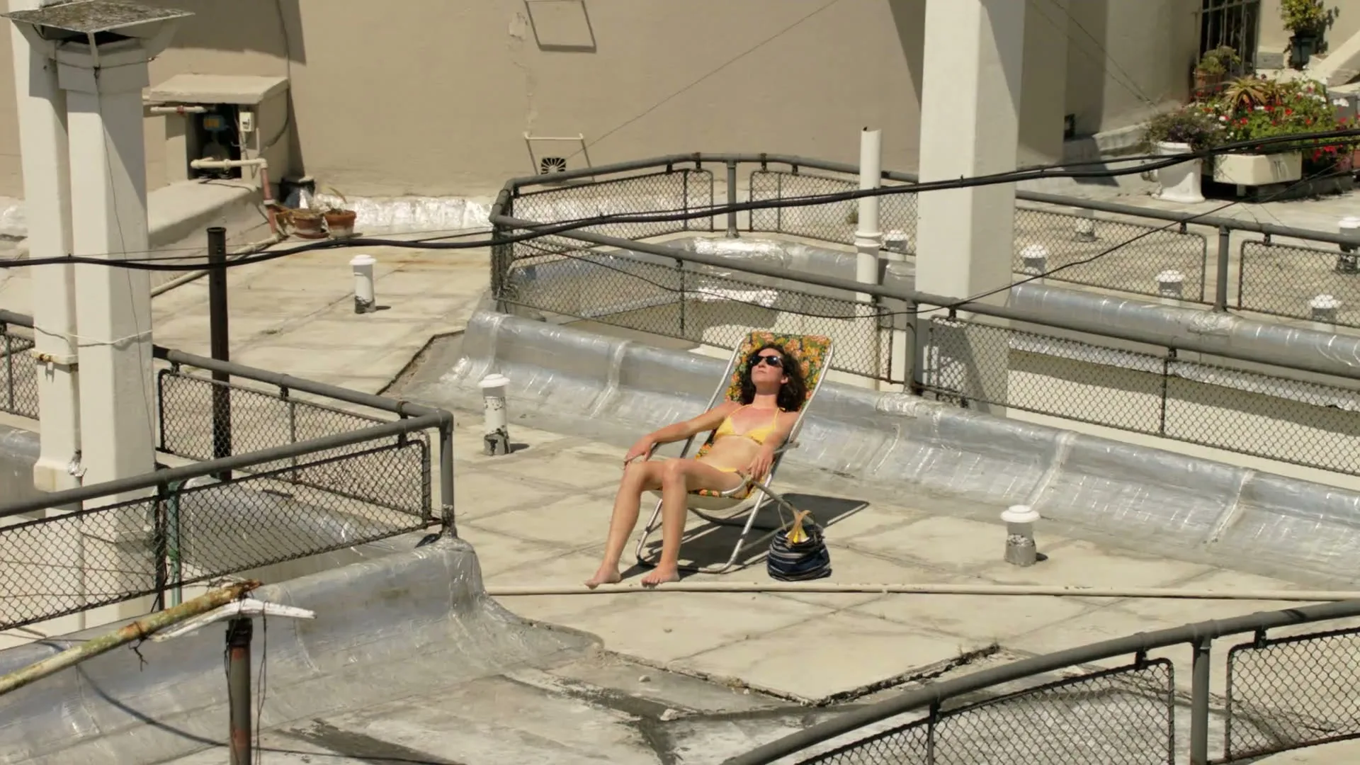 A woman in a yellow bikini and sunglasses sunbathes on a rooftop in a lounge chair, looking relaxed on a sunny day.