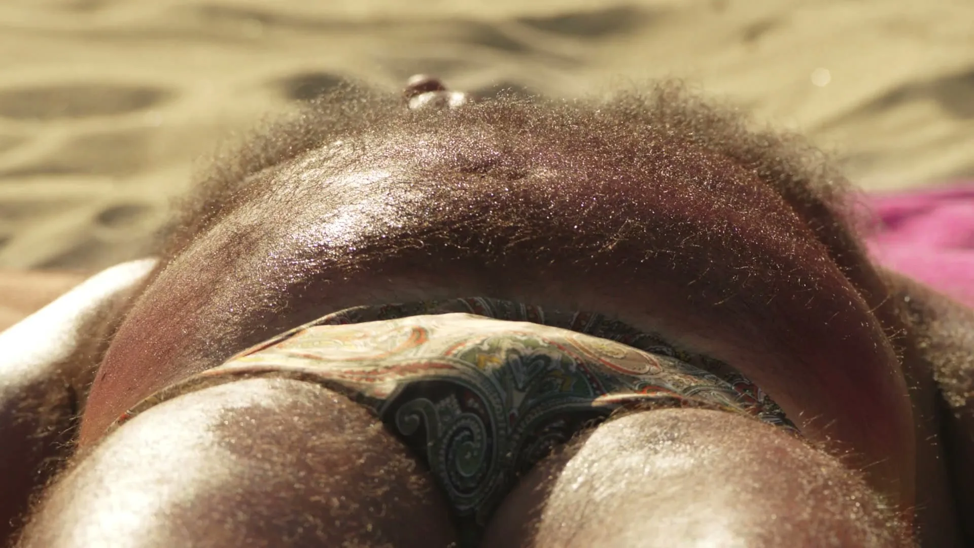 A hairy person with dark skin and a patterned swimsuit lies on a sandy beach, glistening with sweat under the sun.