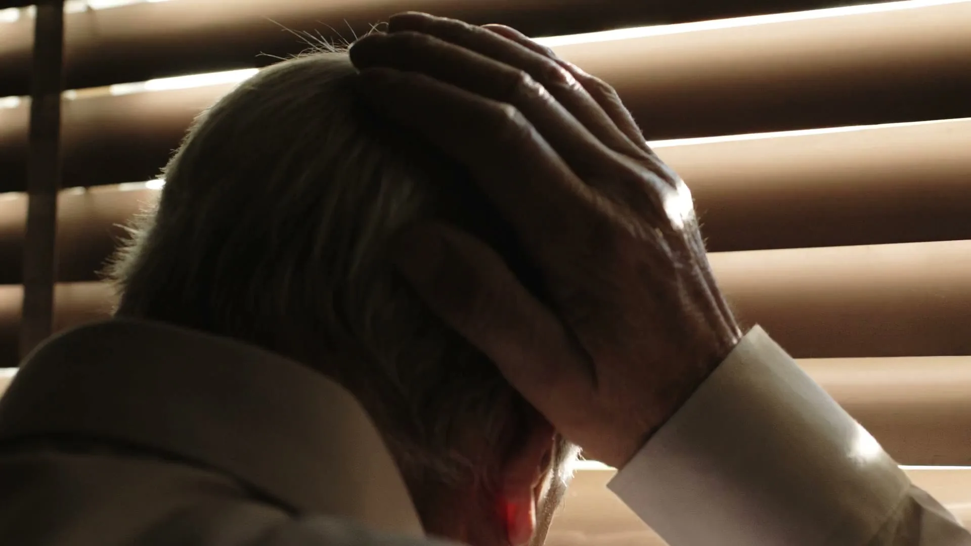 An older man with white hair, wearing a white shirt, holds his head in his hands, looking down in despair, silhouetted against window blinds.