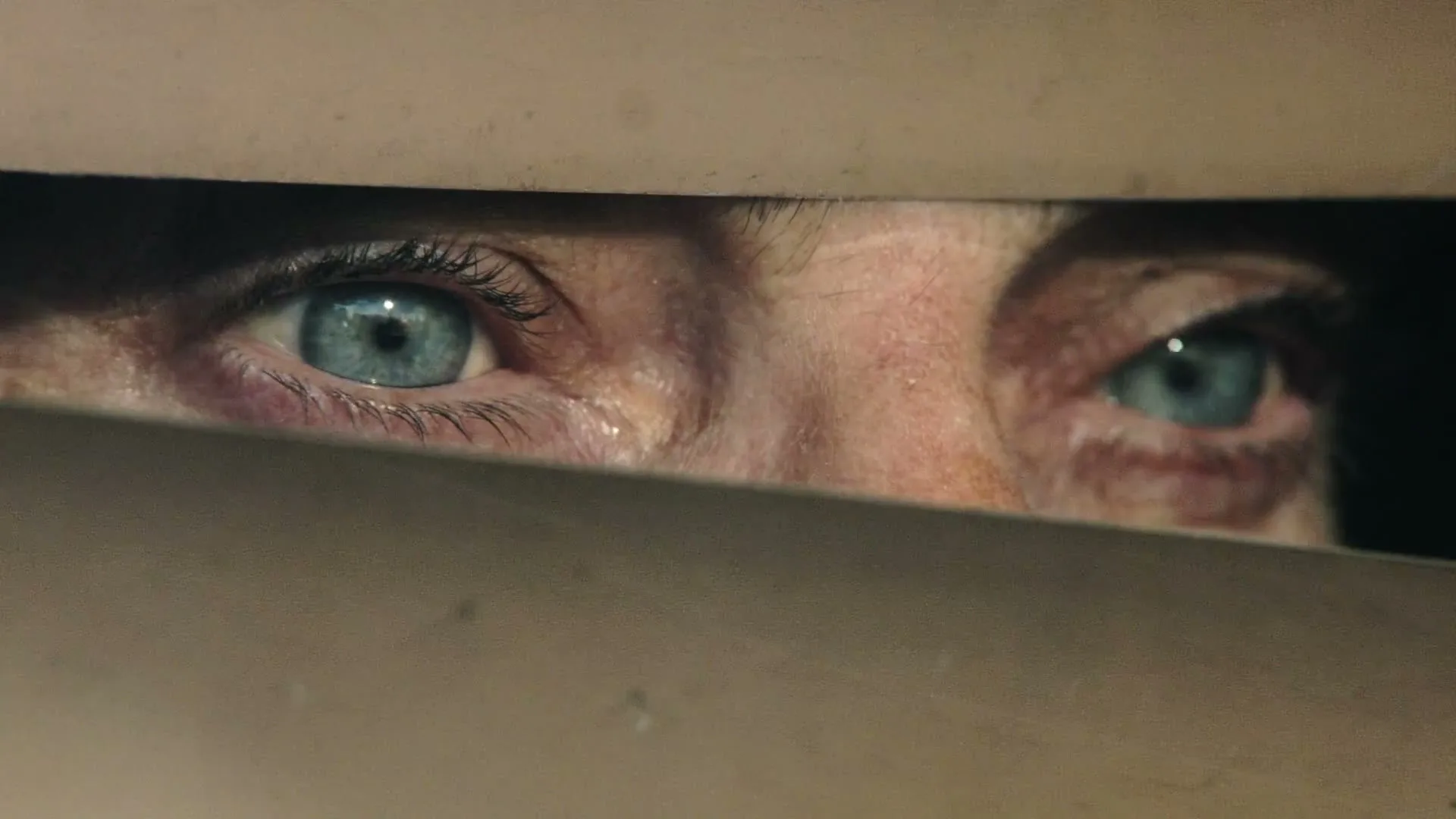 A person's blue eyes peer intensely through a narrow horizontal slit, their face partially obscured by a beige surface.
