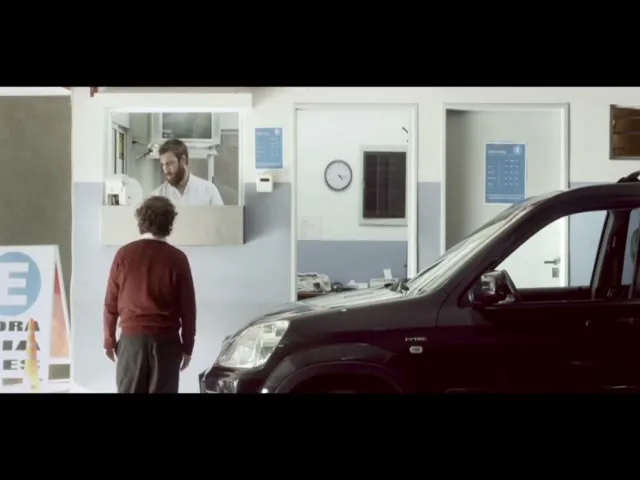 A man in a white coat looks at a man in a red sweater who stands next to a black car at a service window.