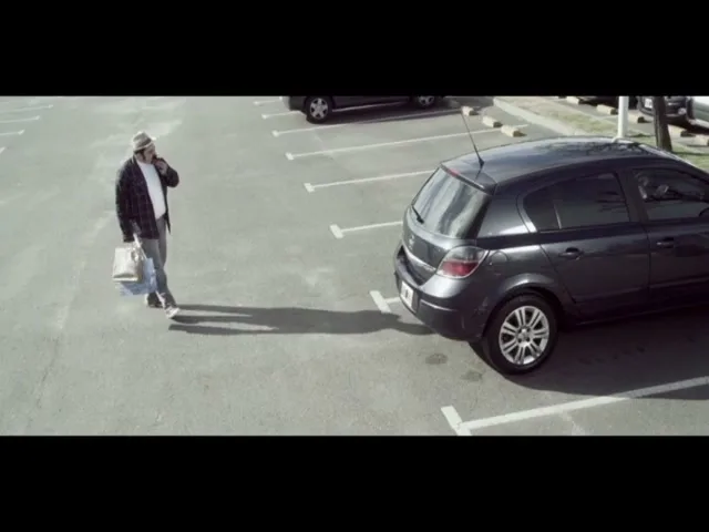 A man in a hat and plaid shirt walks across a sunny parking lot, talking on his phone and carrying a shopping bag, towards a dark grey hatchback car.